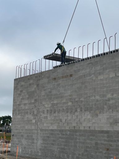 Worker on top of a tall concrete wall, secured by a crane cable at a construction site.