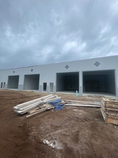 Construction site with unfinished warehouse bays, dirt foreground, stacked lumber, and cloudy sky