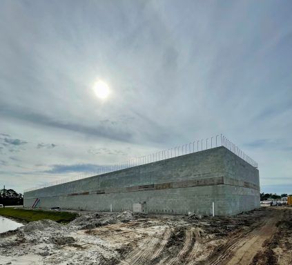 Large gray concrete building under construction on a muddy site beneath a cloudy sky.