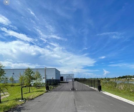 Paved driveway leading to a black gate beside a white industrial building under a bright blue sky