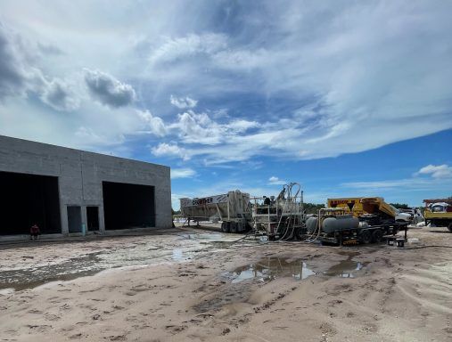 Mud-filled construction yard with damaged vehicles, equipment, and a large industrial building under a cloudy sky