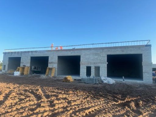 Single-story gray concrete building under construction with three large garage openings and dirt foreground