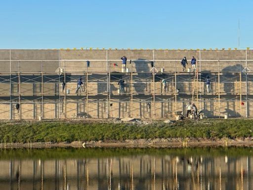 Workers on scaffolding repairing a large beige wall beside water, with reflections below.