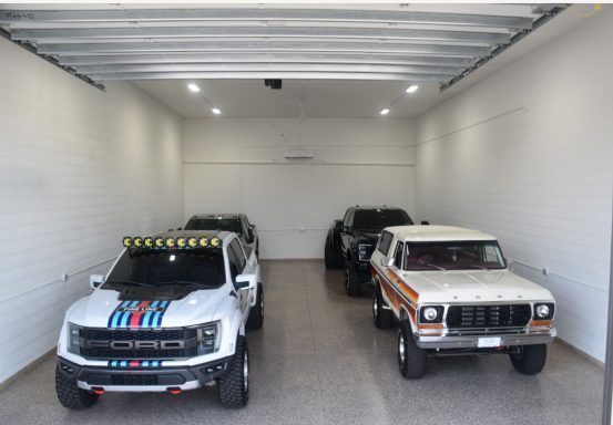 Two off-road trucks parked inside a white garage, one silver with blue stripes and one white with orange stripes.