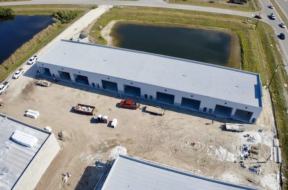Aerial view of a long gray warehouse beside a pond, with parked trucks and a gravel lot