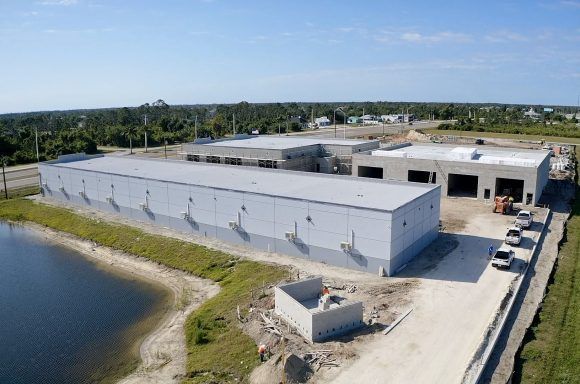 Aerial view of a large industrial building beside water, with a paved yard and surrounding trees.