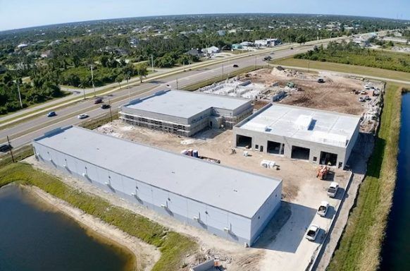 Aerial view of a large industrial building under construction beside a highway, with fields and neighborhood in the distance