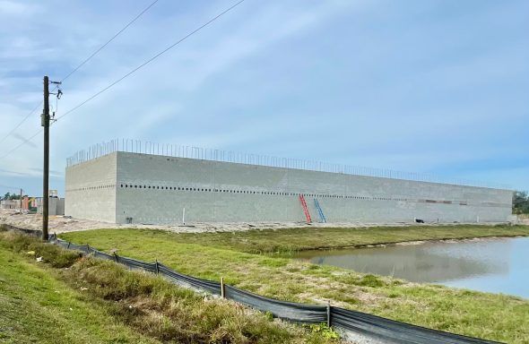 Long white industrial building beside a pond under a blue sky