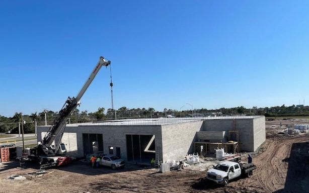 Construction site with crane lifting materials beside a partly built concrete house under a blue sky