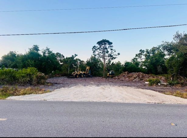 Empty gravel lot beside a road, with trees and a few parked vehicles in the background.