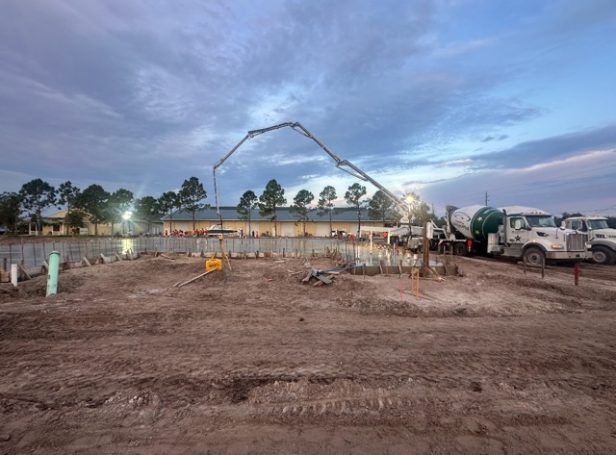 Construction site at dusk with concrete trucks, a large mixer arm, and a row of trees in the background