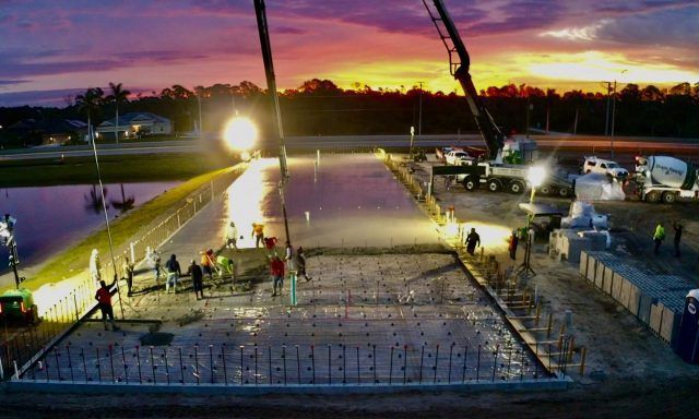 Construction workers pour concrete on a bridge deck at dusk, with cranes and bright work lights overhead