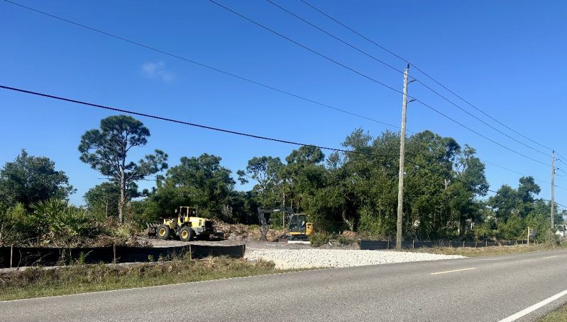 Rural roadside with utility poles, trees, and a few houses under a clear blue sky.