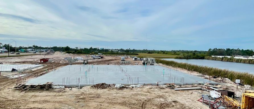 Construction site with a large rectangular concrete foundation beside a pond under a cloudy sky