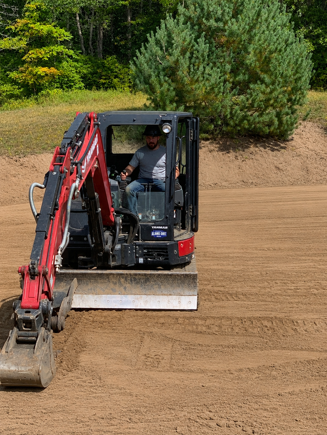 Red mini excavator digging dirt on a hillside with shrubs and trees nearby