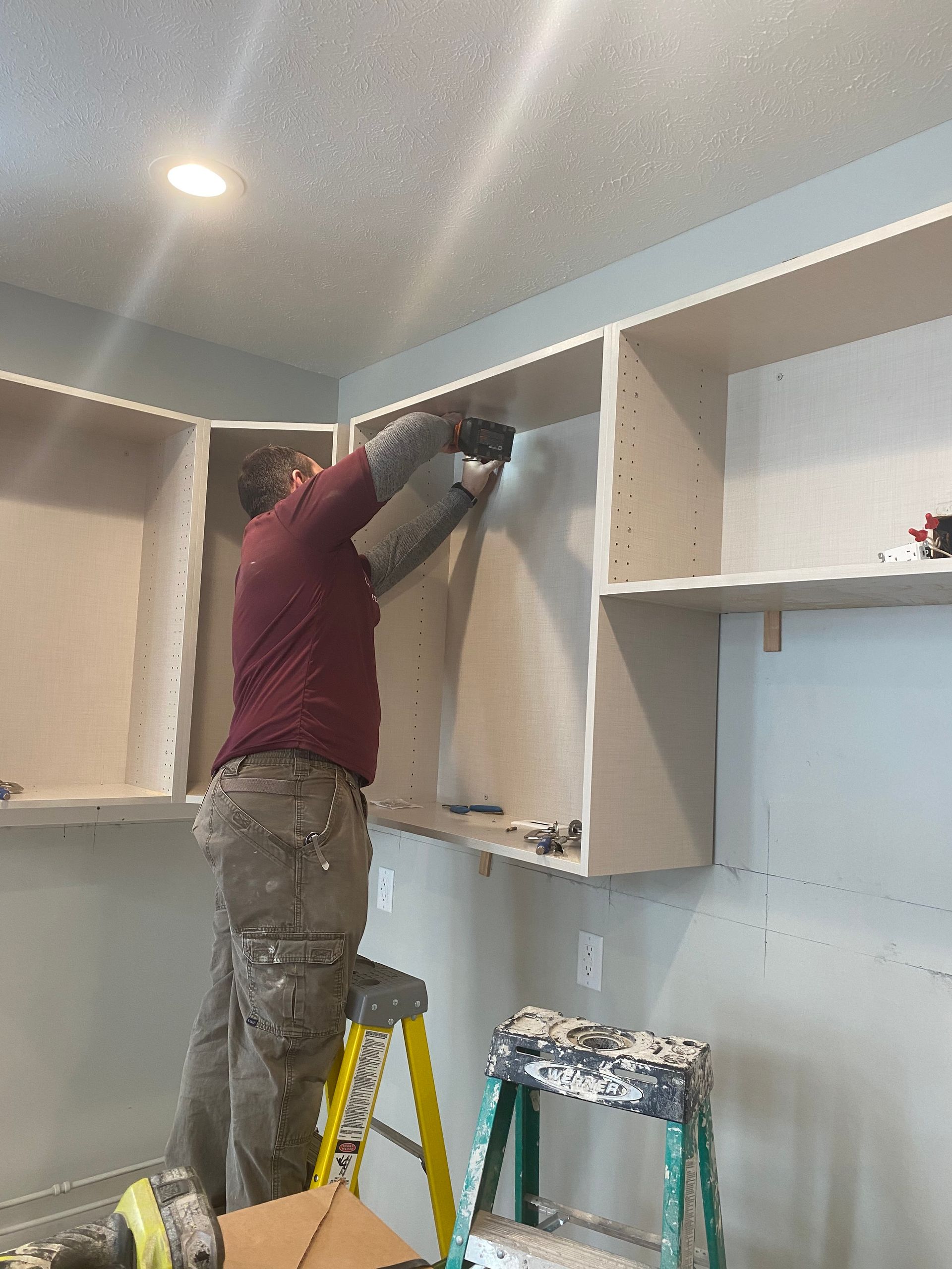Person installing a cabinet in a room, standing on a yellow step ladder with tools nearby.