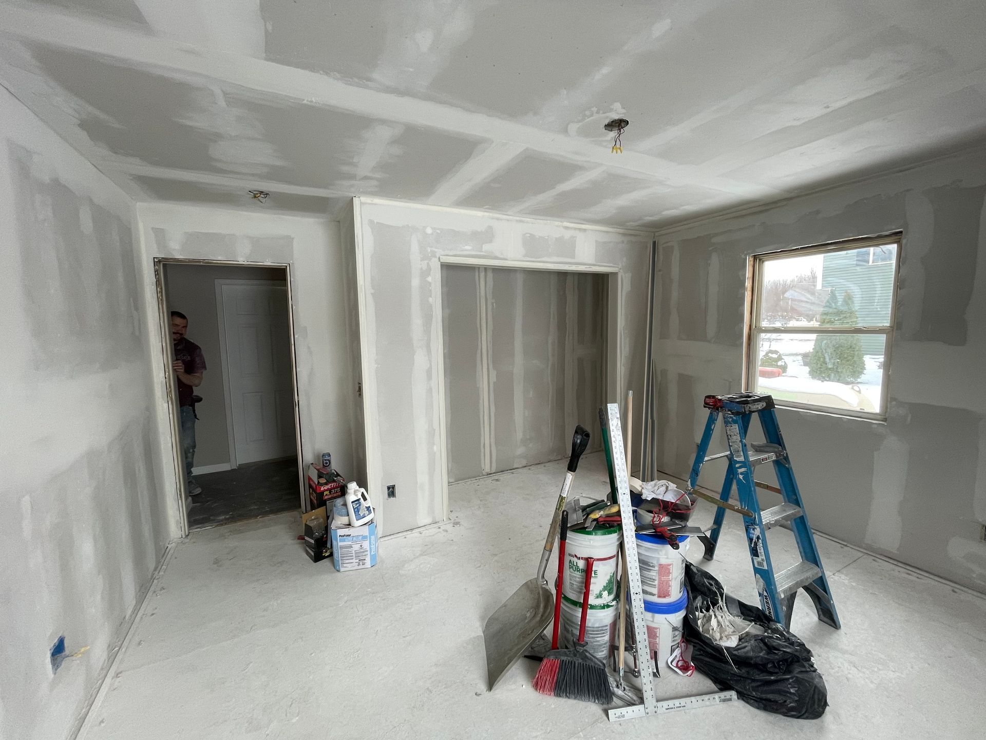 Unfinished room with drywall, paint supplies, ladders, and a window letting in daylight