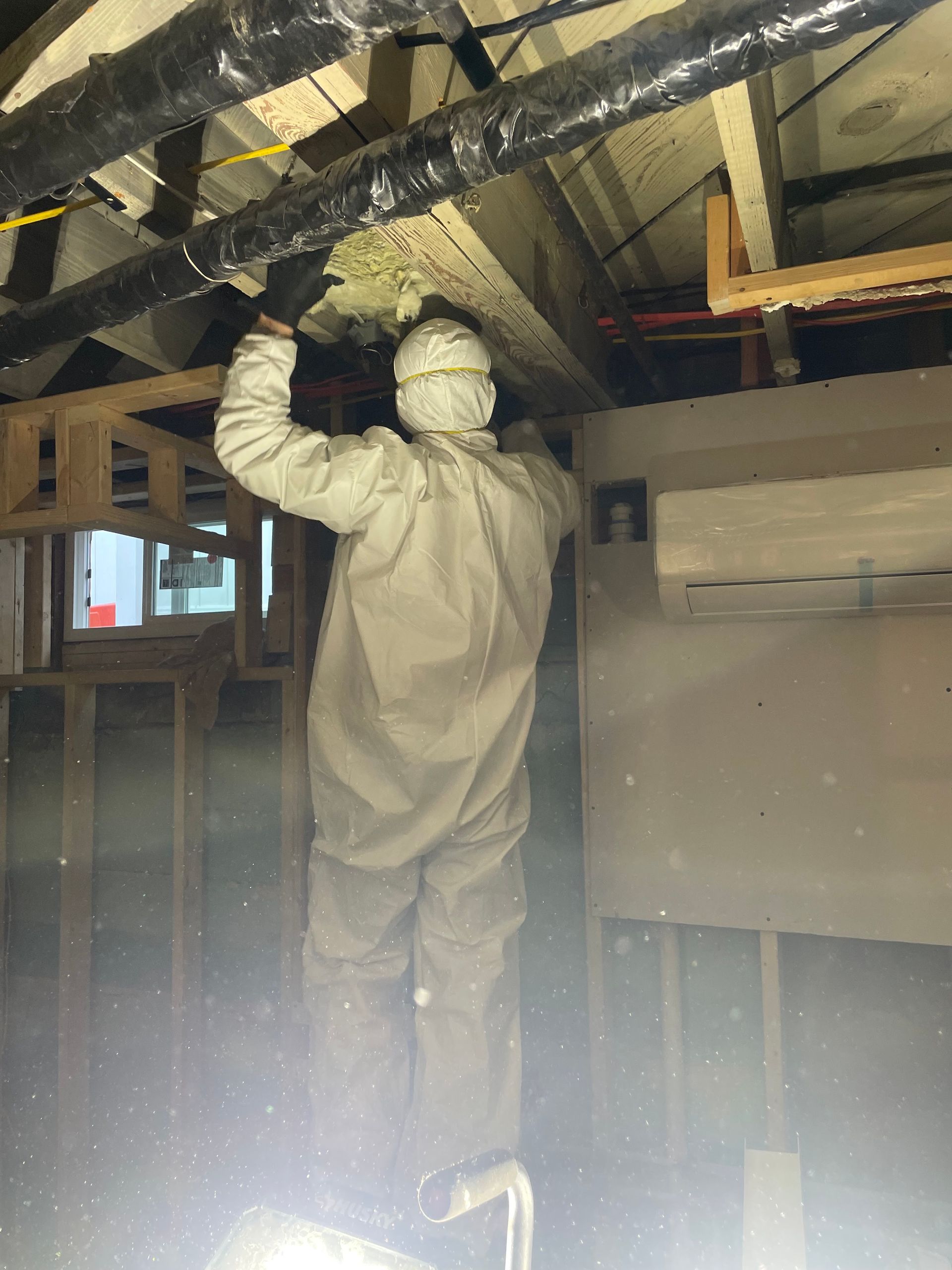 Worker in white coveralls installing insulation in an attic with exposed beams.