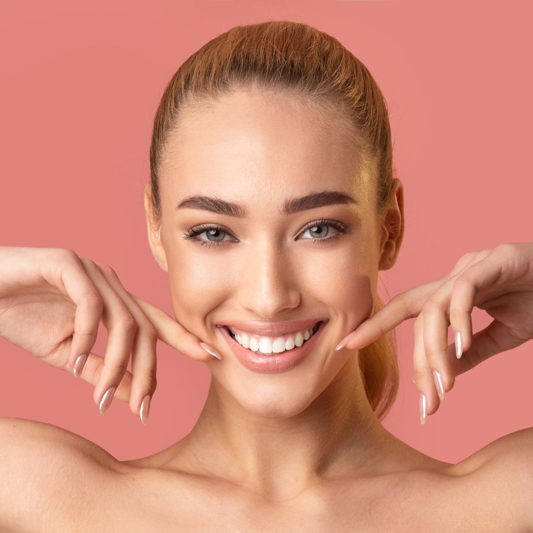 A person with light-brown hair and a radiant smile, framing their face with both hands against a soft, solid pink background.