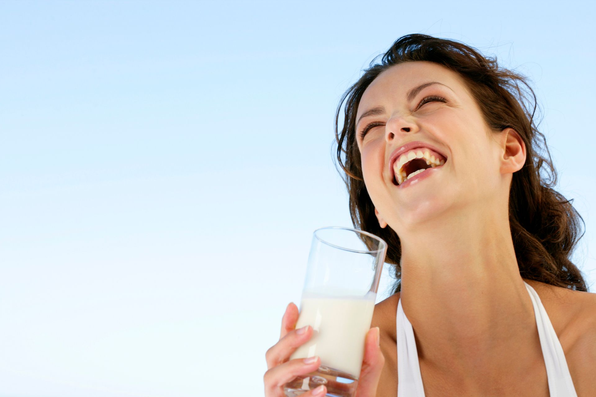 A person laughing while holding a glass of milk against a clear blue sky background.