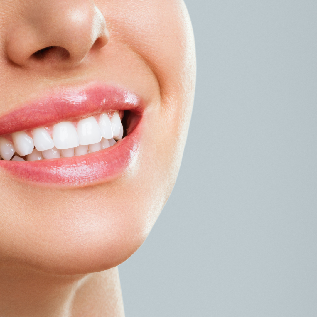 A close-up of a person smiling, showing straight, white teeth and pink lips against a plain, light grey background.