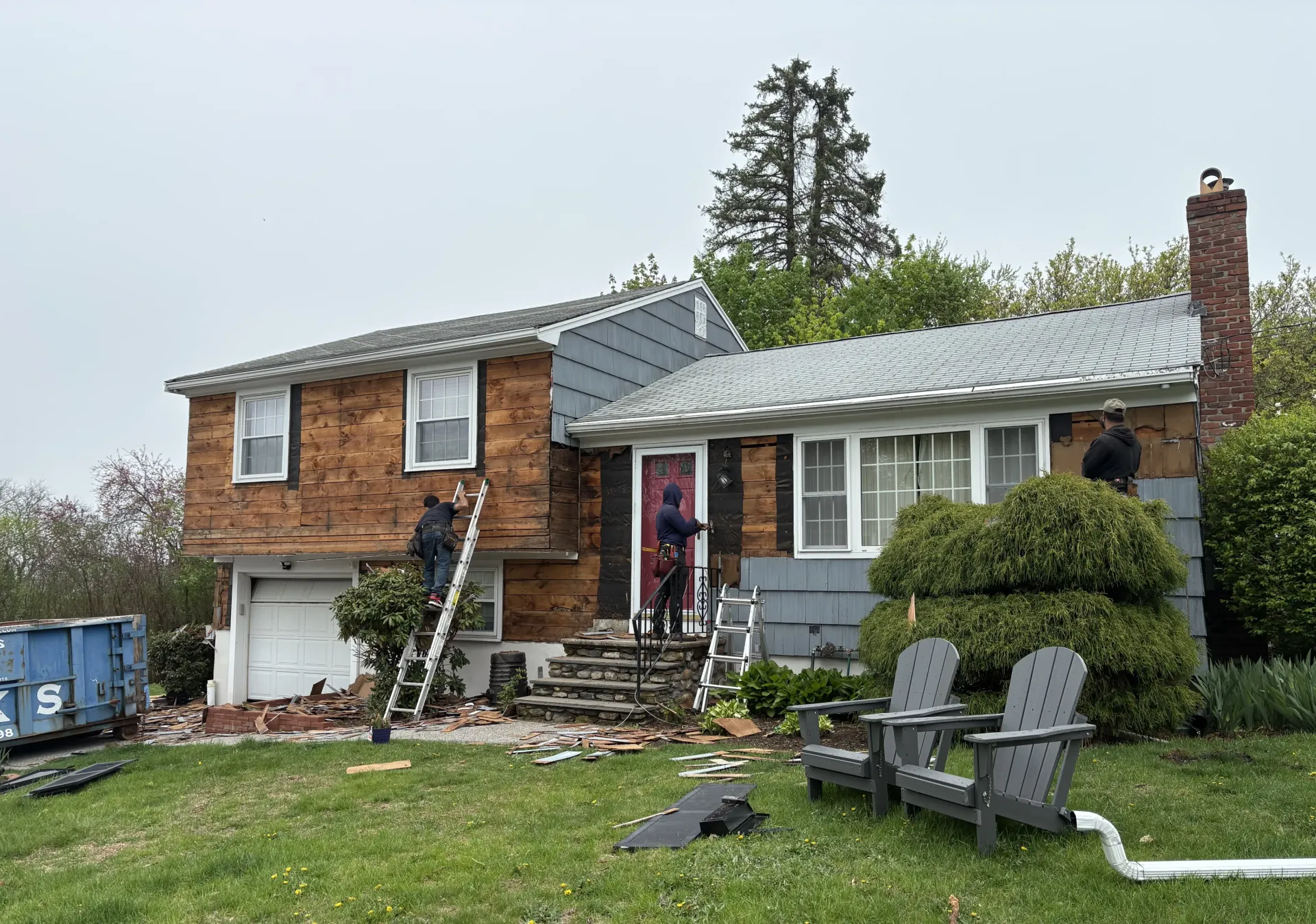 House under renovation with workers on ladders, brick siding removed. Gray siding and green lawn.