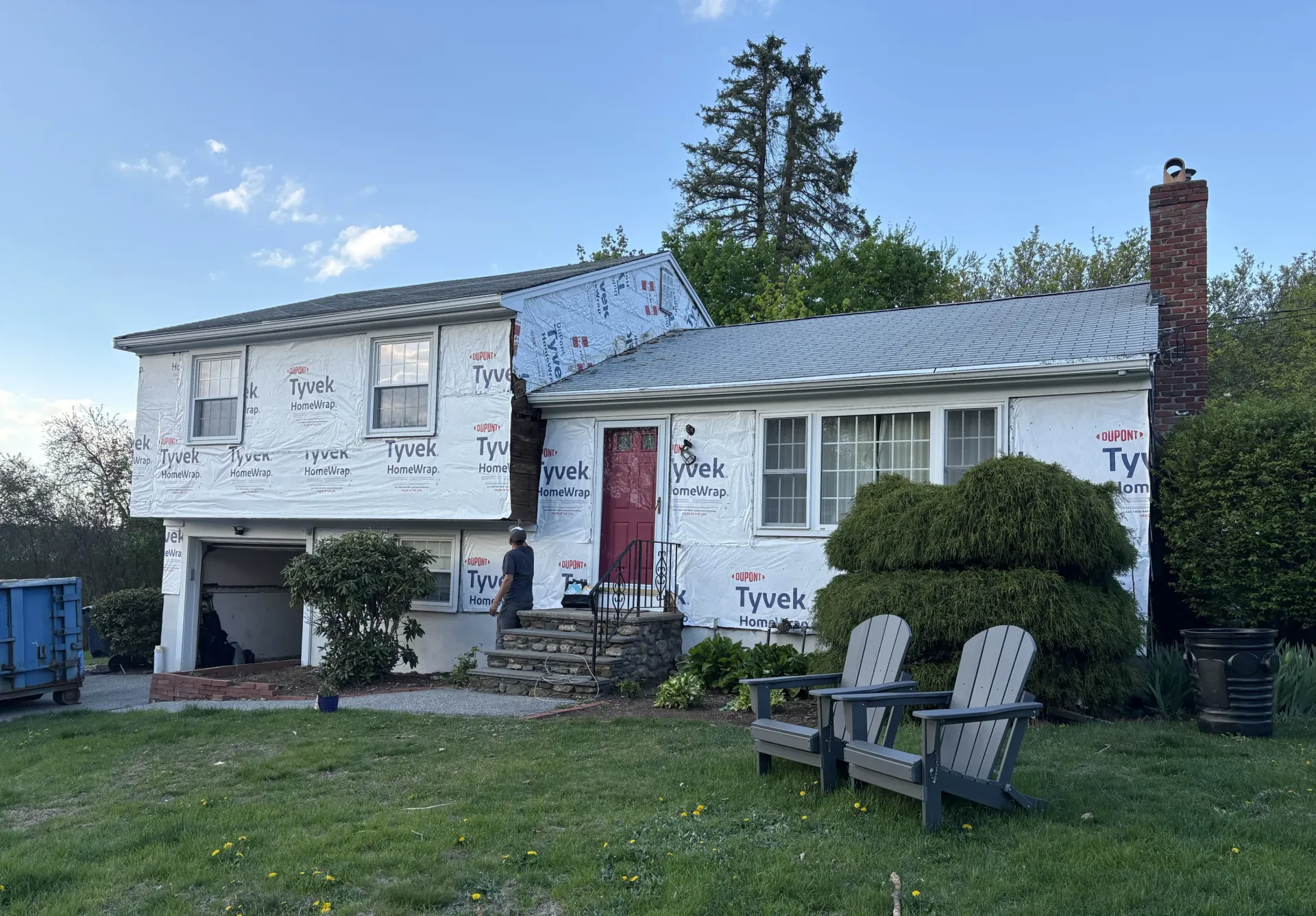 House under construction, covered in white Tyvek wrap, red door, chimney, and two wooden chairs in yard.