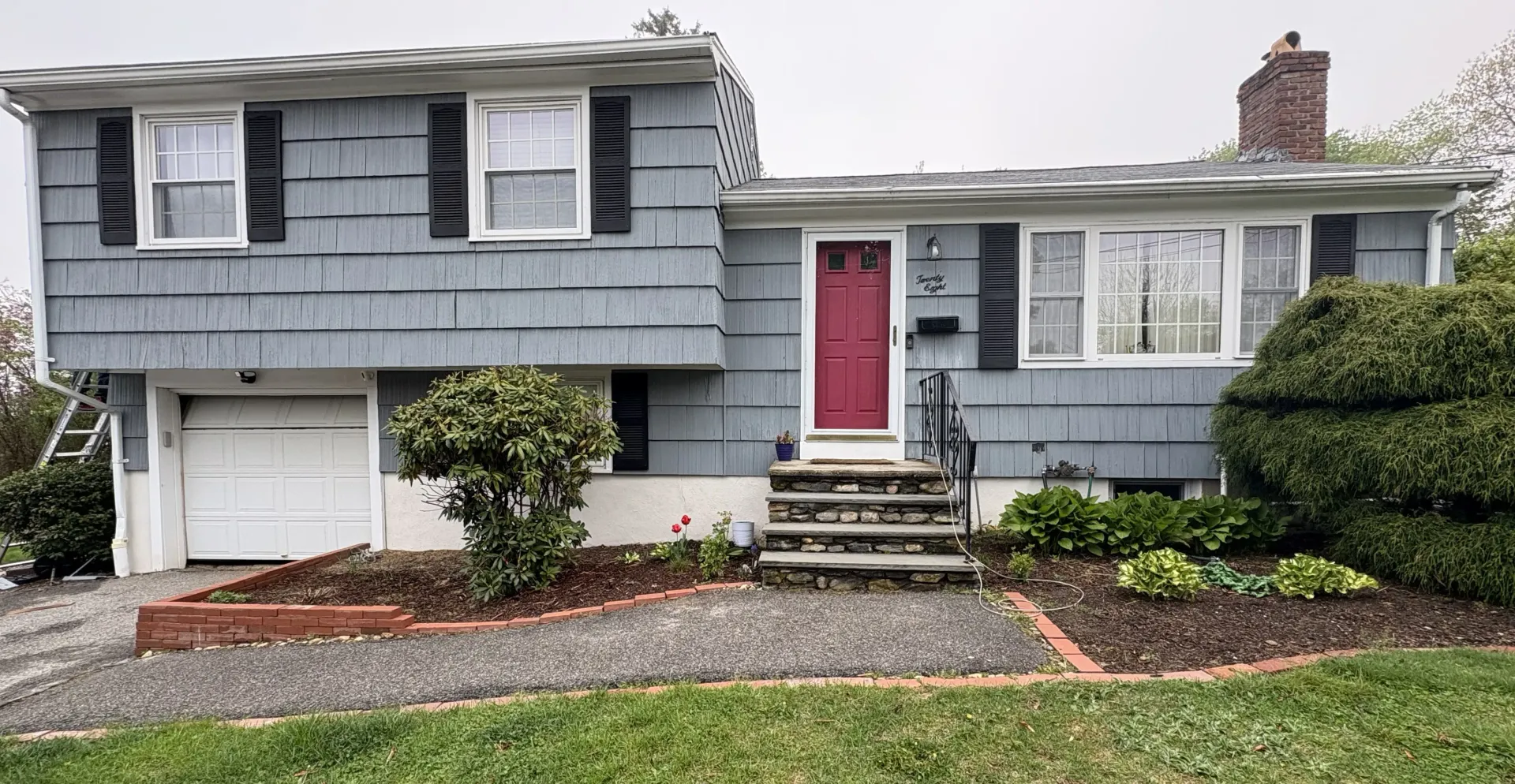 Split-level house with gray siding, a red door, and a small front yard with bushes.