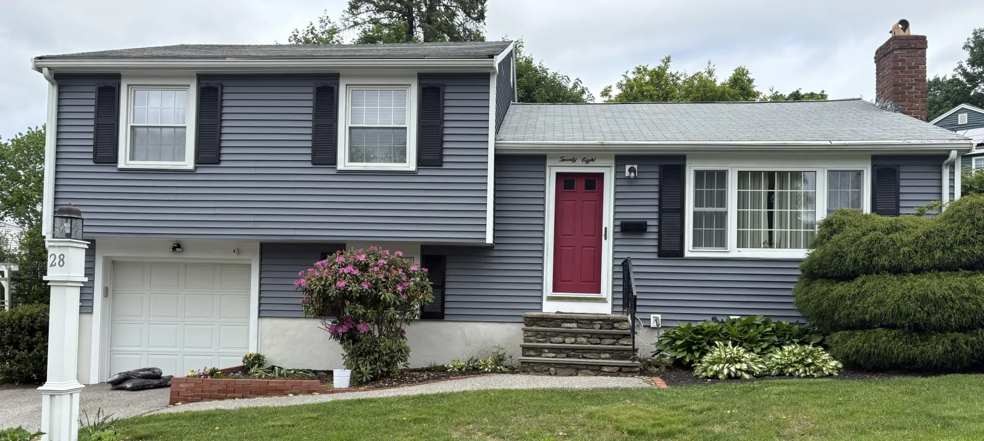 Two-story gray house with a red door, black shutters, and a garage. Green grass and bushes in front.