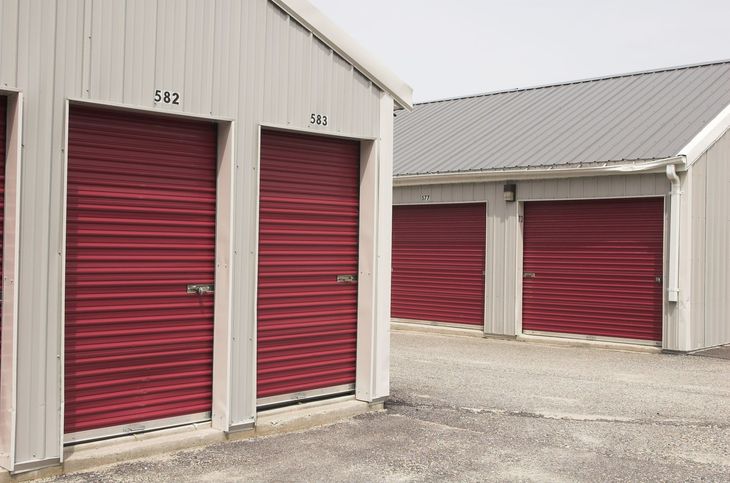 Rows of red storage unit doors in a gravel lot, under white metal buildings.