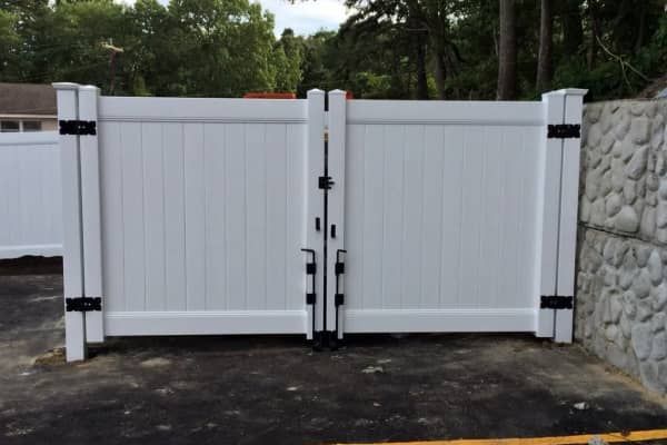 White vinyl double gate, black hardware, set in a driveway. Stone retaining wall on the right.