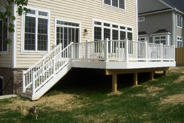 White deck with stairs, attached to a light beige house on a grassy hill, surrounded by trees.