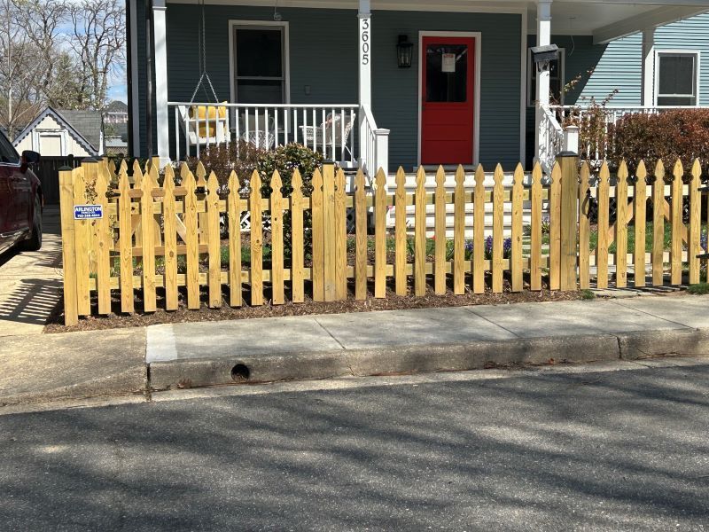 Yellow picket fence in front of a blue house with a red door, on a sunny day.