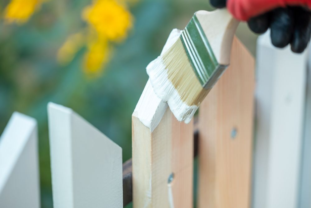 Painting a white picket fence with a brush, person's gloved hand visible.