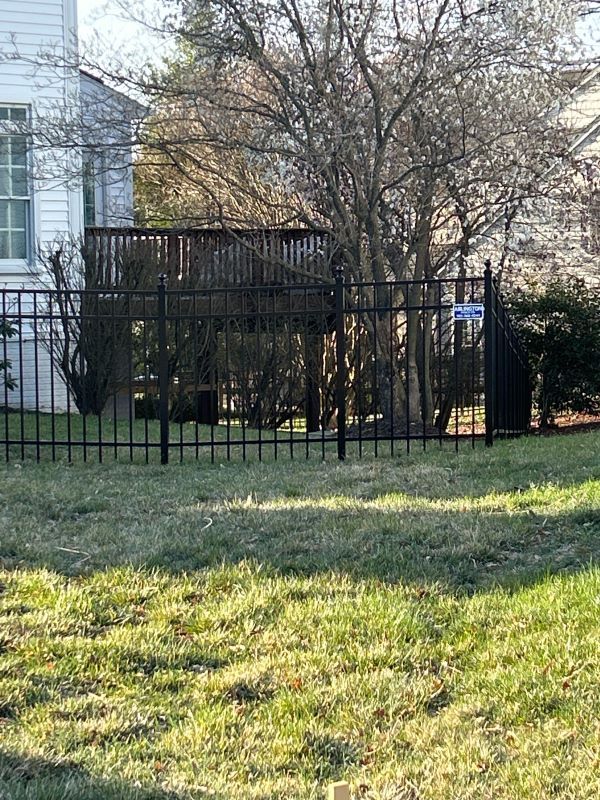 Black fence in front of a yard with a small deck and a tree. A house is in the background.