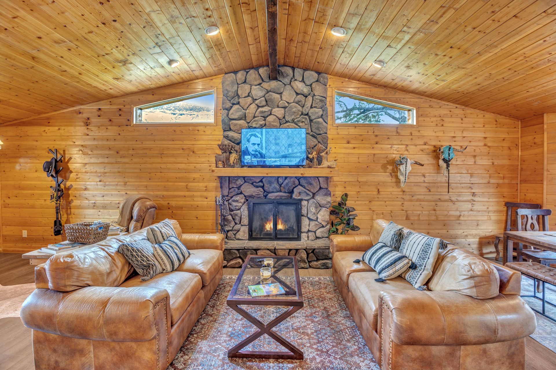 Cozy log cabin living room with stone fireplace, leather sofas, and wood-paneled vaulted ceiling