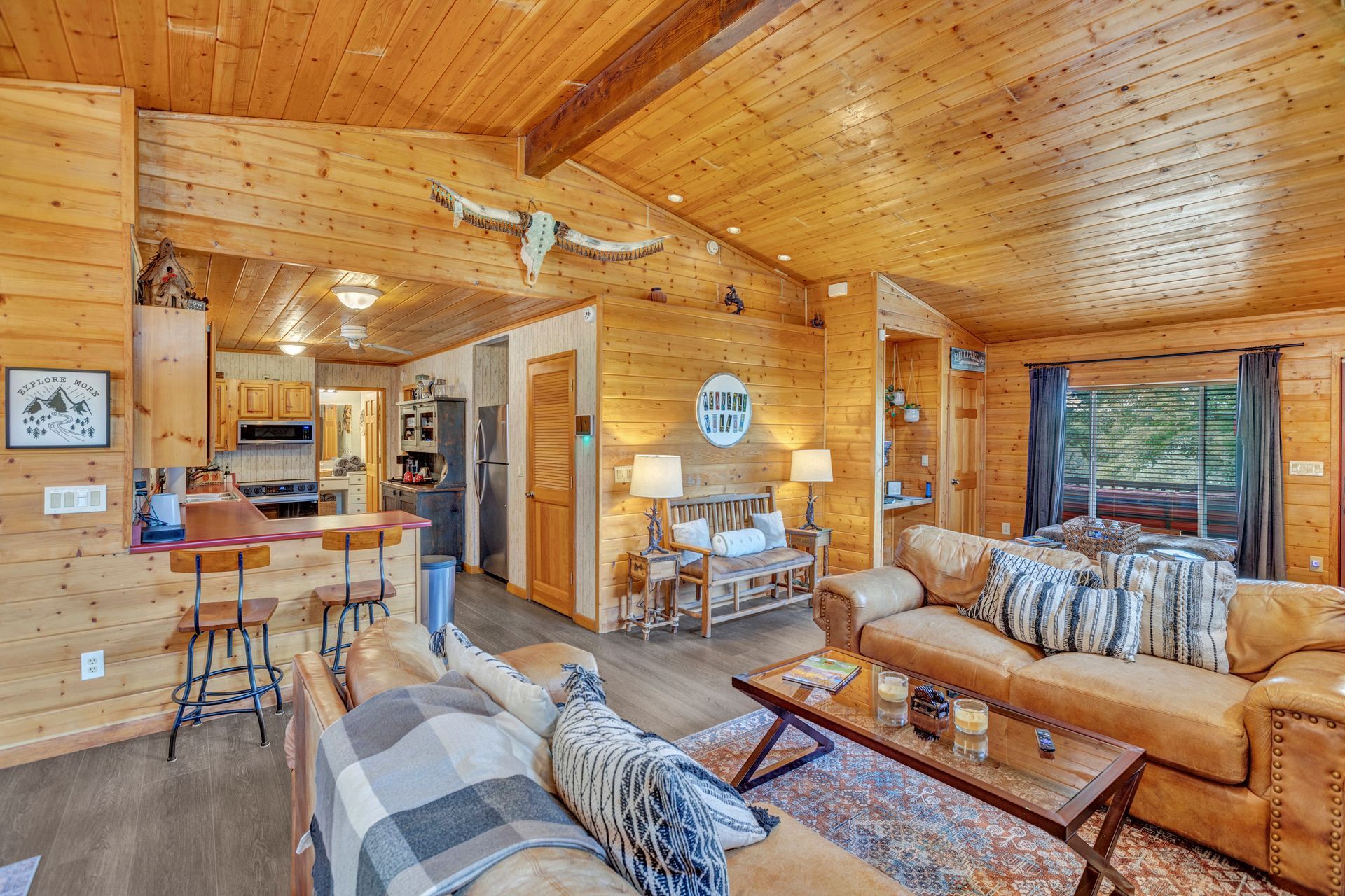 Cozy wood-paneled cabin living room with tan sofas, plaid accents, bar stools, and vaulted ceiling