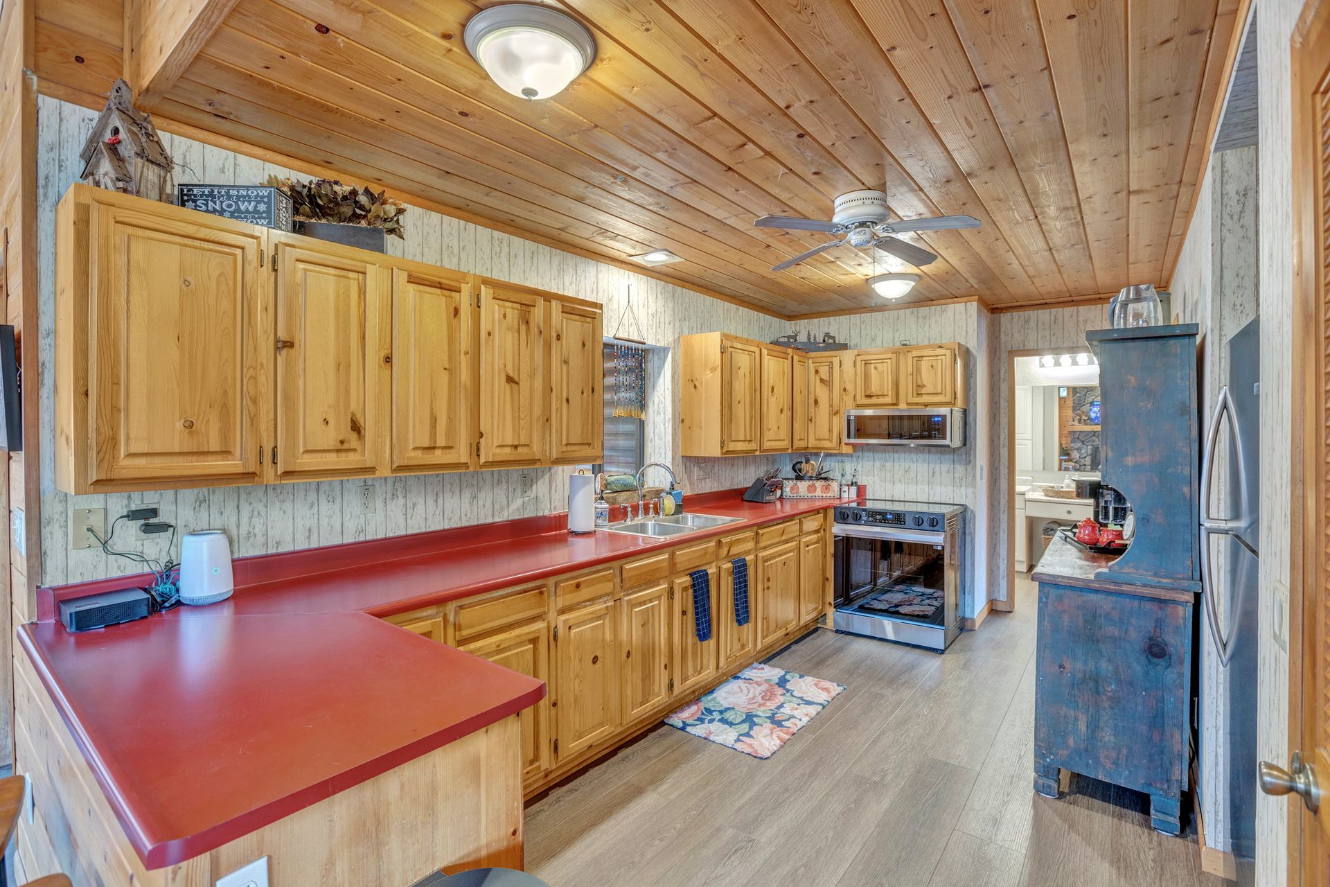 Rustic kitchen with wood cabinets, red countertops, wood-paneled ceiling, and stainless stove and fridge.