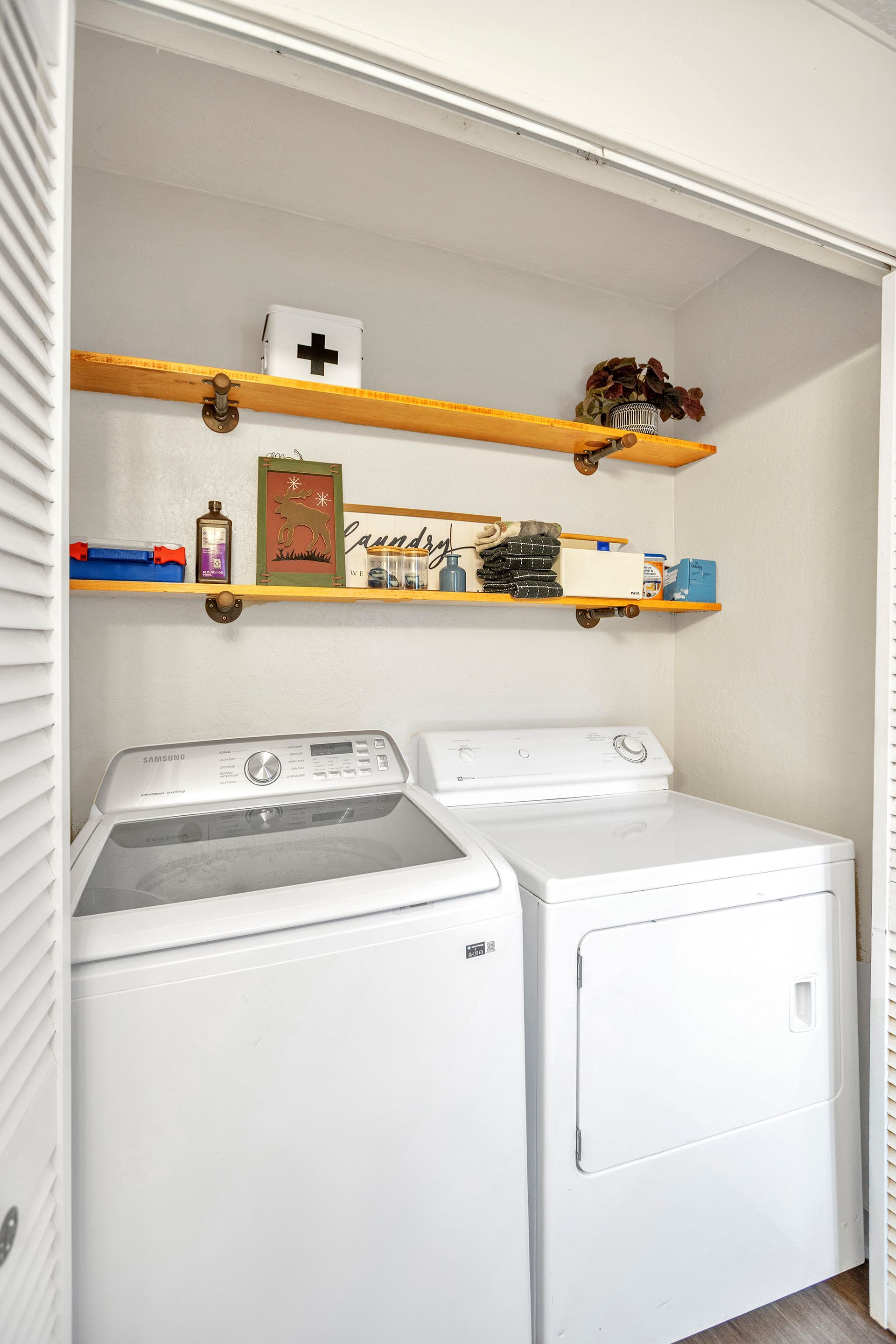 Laundry closet with a white washer and dryer, shelves above holding cleaning supplies and decor.