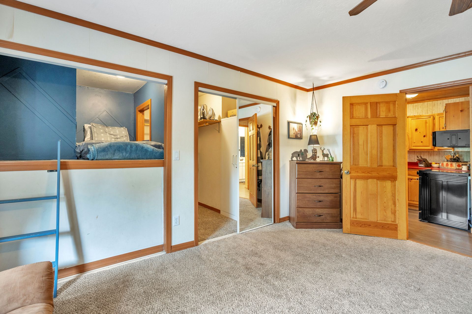 Bright bedroom with carpet, open closet, dresser, hallway, and kitchen visible through a doorway.