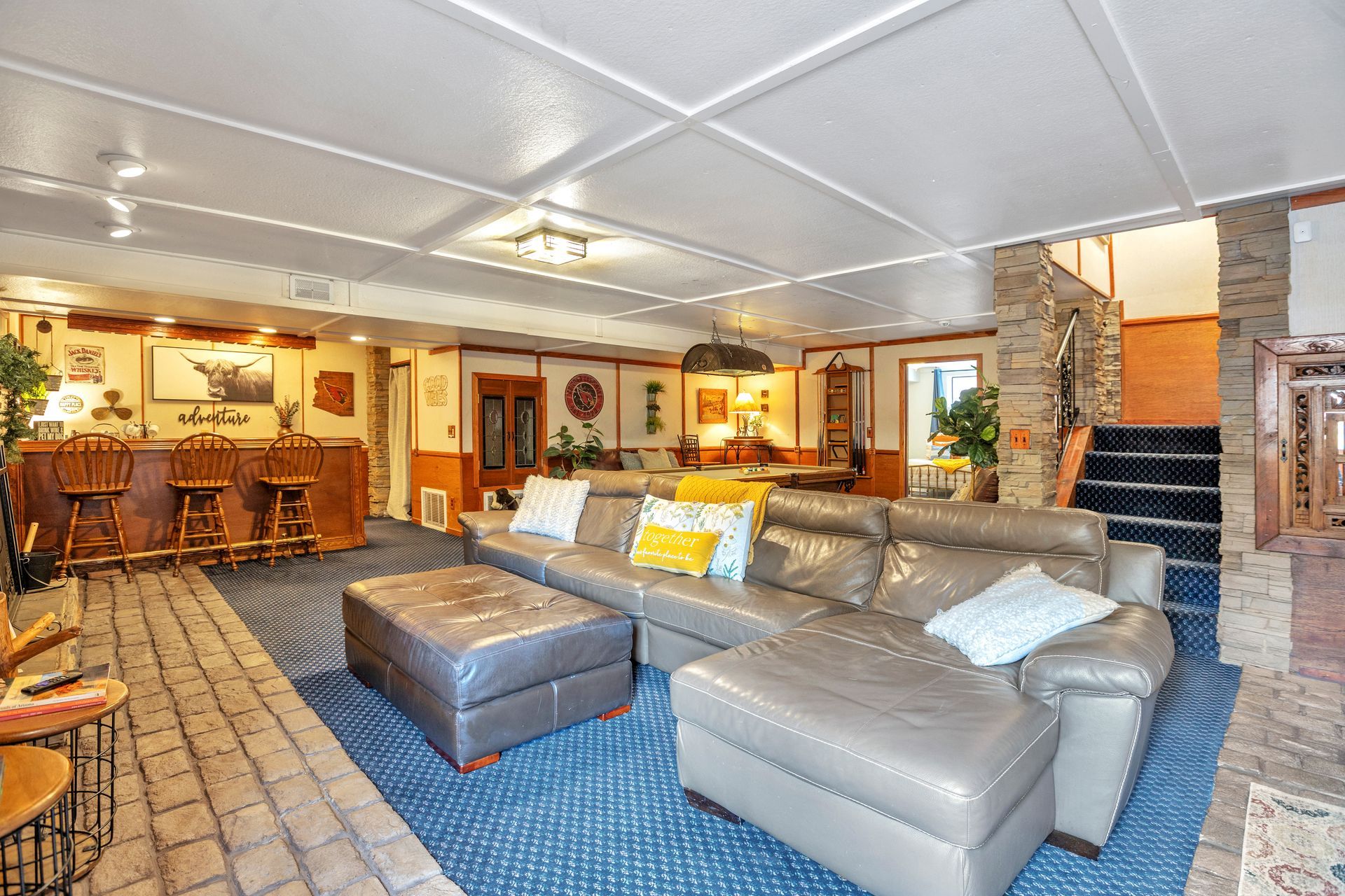 Spacious living room with gray sectional, ottoman, stone fireplace, and open dining area with wood tables