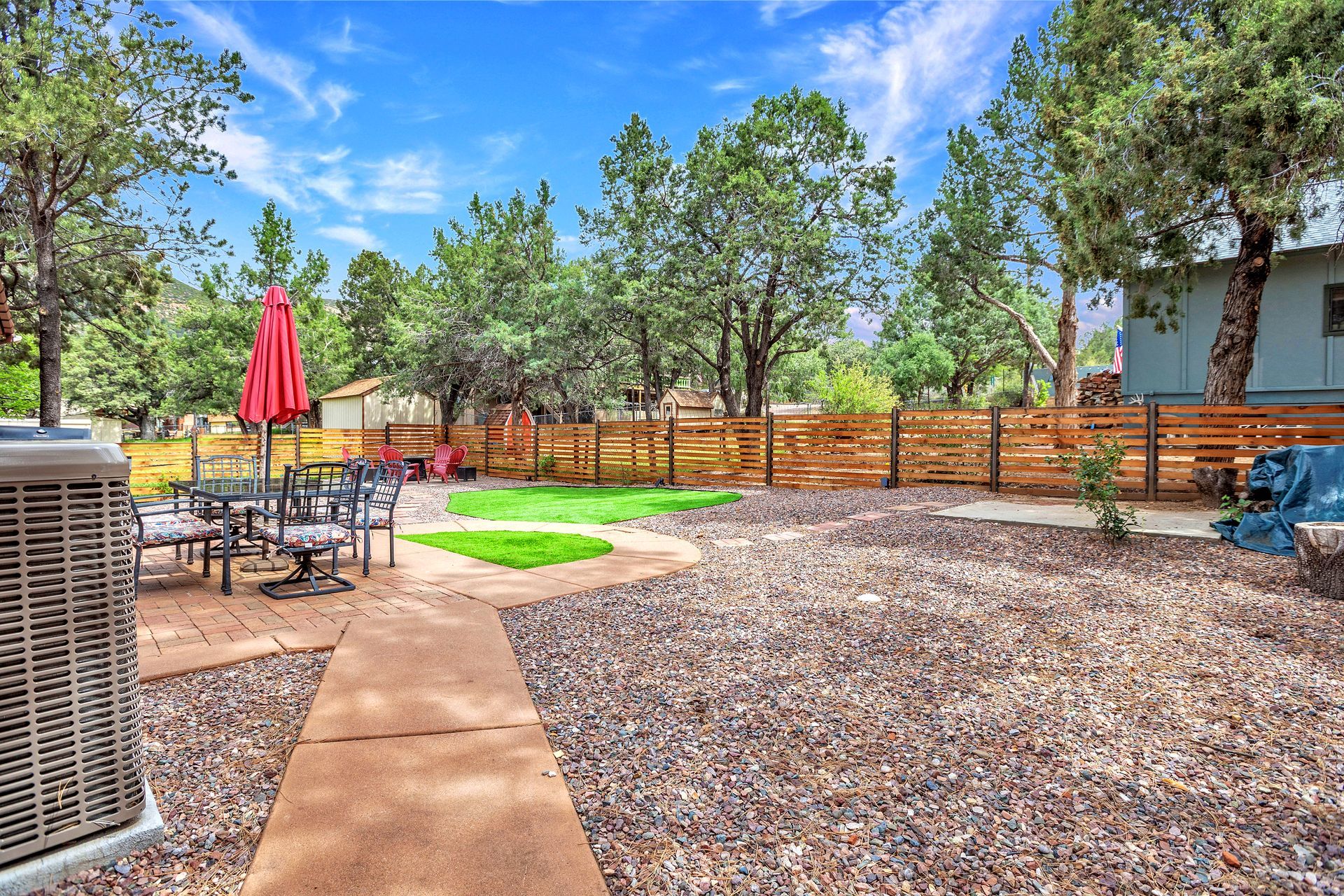 Backyard patio with stone path, red umbrella, grill, and wooden fence under a bright blue sky