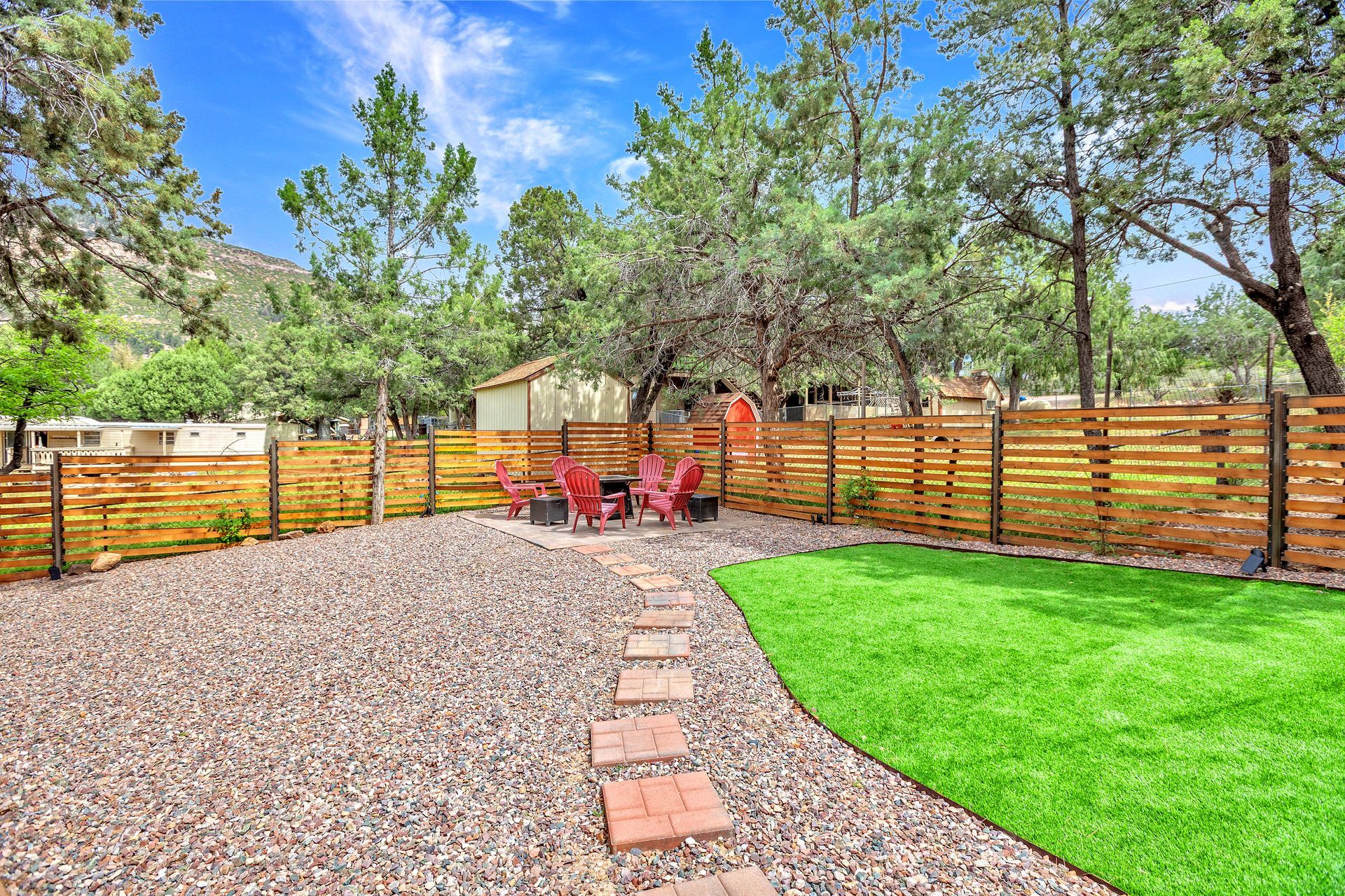 Backyard patio with brick path, red chairs, gravel, green lawn, wooden fence, and trees under a blue sky