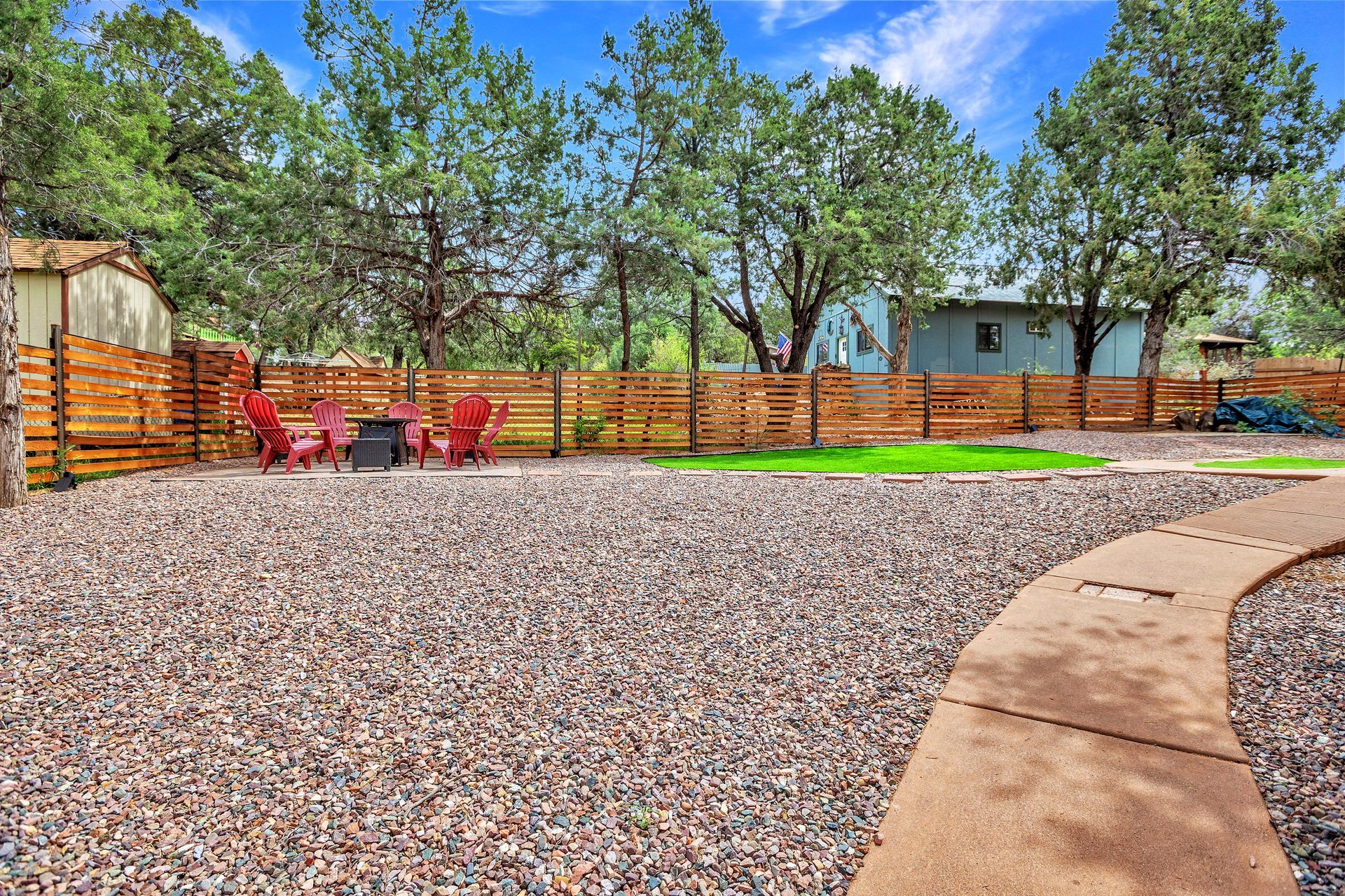 Gravel backyard patio with red chairs, wooden fence, trees, and a small shed under a blue sky