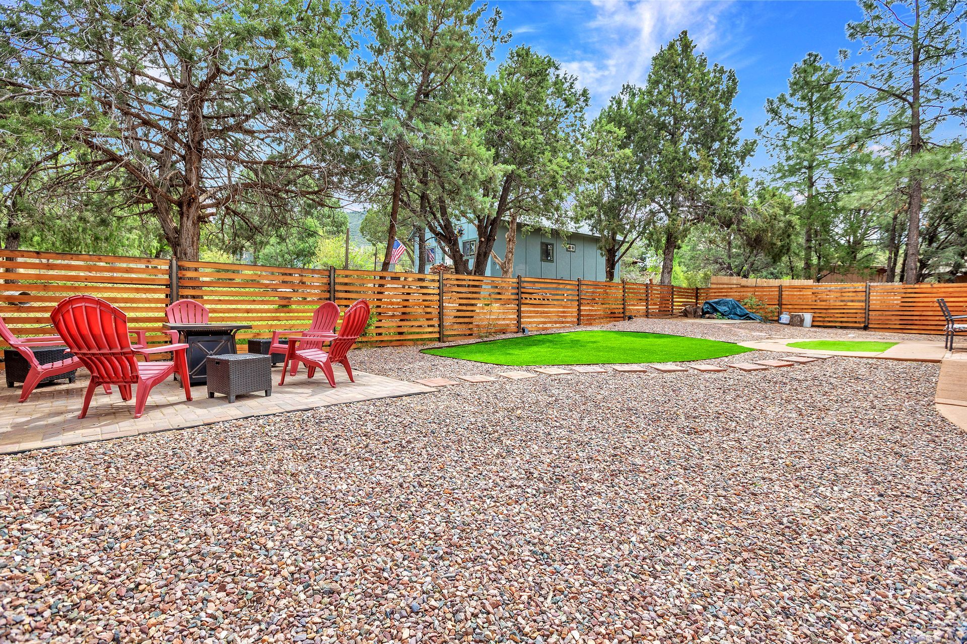 Backyard patio with red chairs, table, wooden fence, trees, and gravel ground
