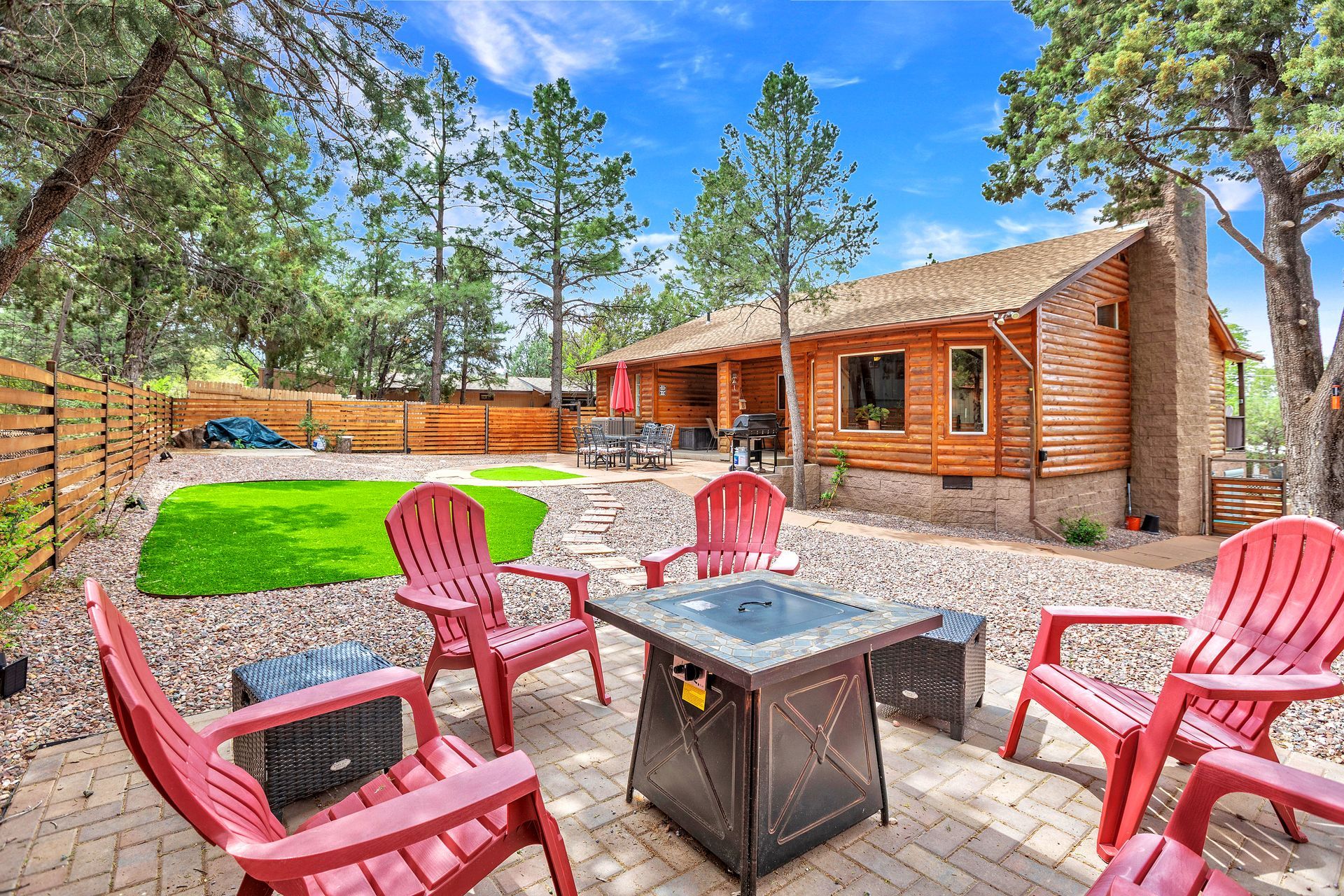 Backyard patio with red chairs around a fire pit beside a log cabin and grassy yard
