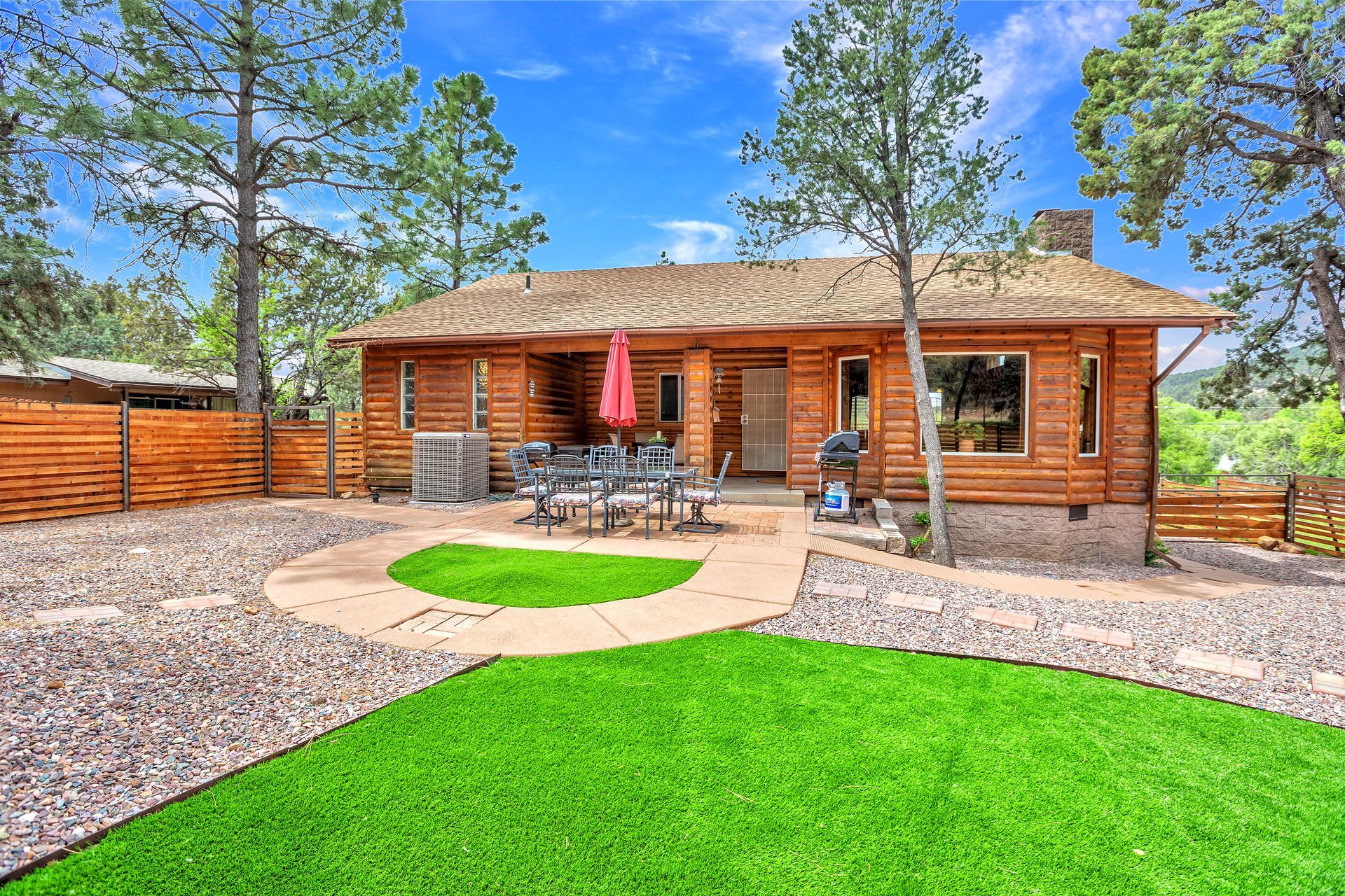 Wooden cabin with patio, red umbrella, stone path, and bright green lawn in a wooded yard.