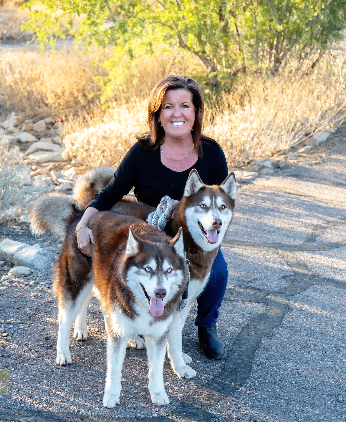 Woman kneeling behind two dogs on a sunny road, both dogs facing forward.