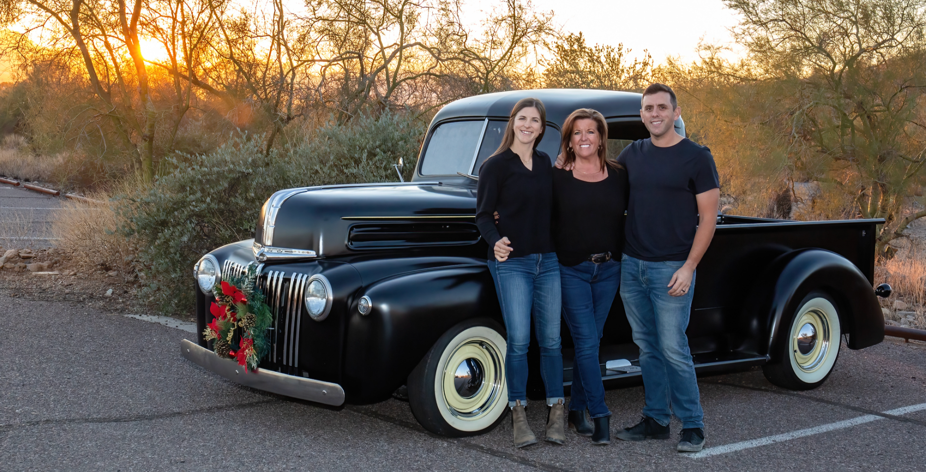 Three people pose beside a black vintage pickup truck at sunset.