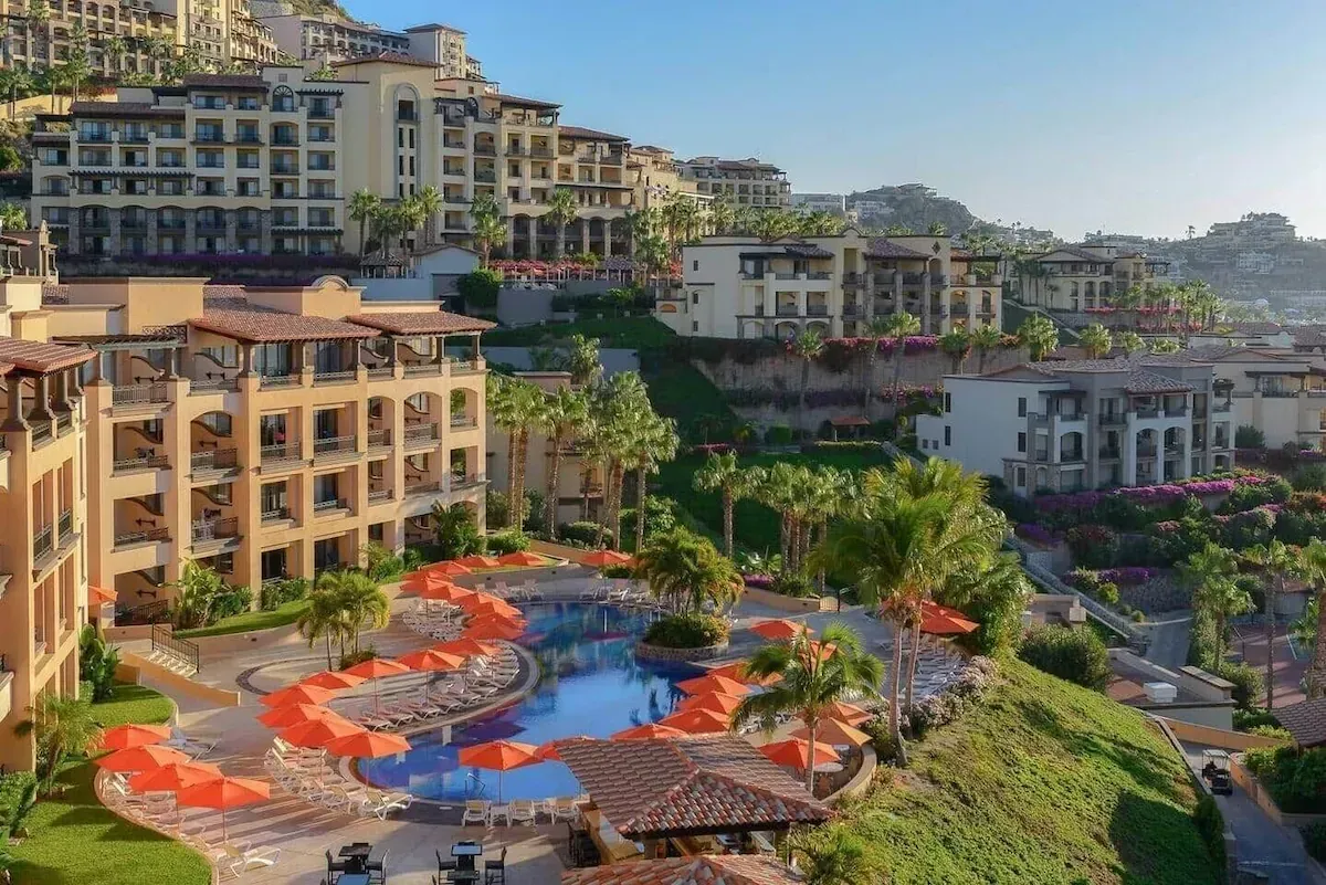 Sunlit hillside resort with multi-level buildings, palm trees, and a blue pool with orange lounge chairs